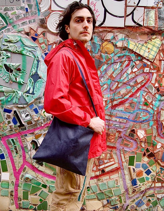 Male model holds navy blue waxed canvas tote bag while standing in an alley of mosiac tile artwork.