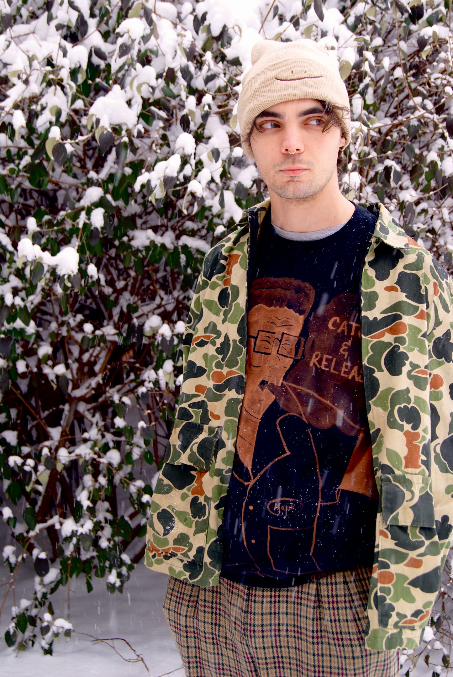 Male model wearing a vintage camouflage zip-up jacket over a back tee with plaid pants, standing in the snow.